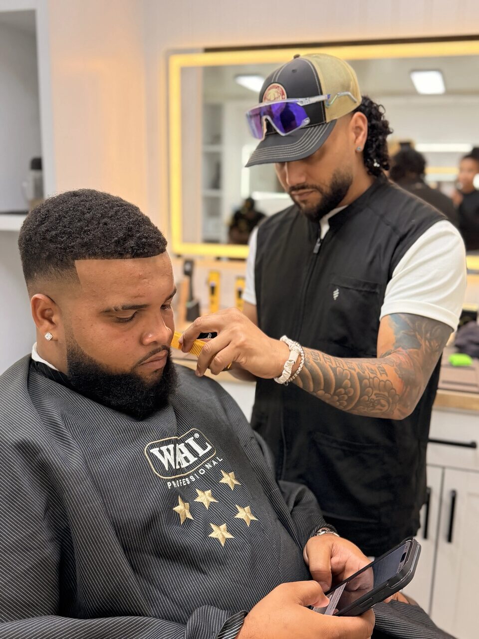 A barber detailing a client's beard beside a lit station mirror